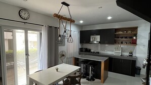 A modern kitchen with a dining area, featuring a white table, wooden chairs, and a unique ceiling light fixture. - 2BR Home (corner lot). With Lanai, 100 Mbps Internet and Netflix. (Balanga)