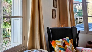A room with a view of a garden and buildings, a chair with a colorful pillow, and a small table with a magazine. - La Forge - Historical Apartment in Sarlat (Sarlat-la-Canéda)