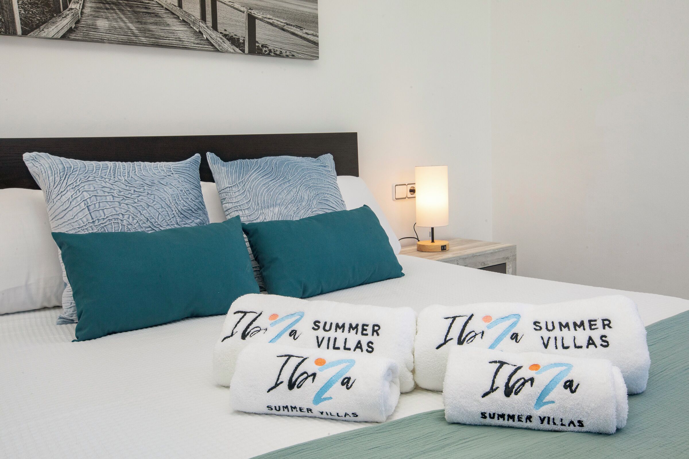 A neatly made bed with teal pillows, a white headboard, and two towels with 'Ita' and 'SUMMER VILLAS' printed on them.