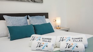 Una cama bien hecha con almohadas color turquesa, un cabecero blanco y dos toallas con la inscripción 'Ita' y 'SUMMER VILLAS'.