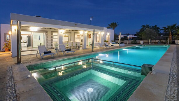 Un área de piscina moderna con un salón con paredes de vidrio, una piscina cristalina y un edificio bien iluminado con techo plano.