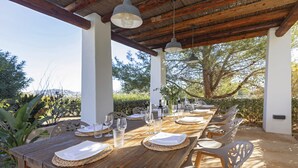 An outdoor dining area with a wooden table set for six, surrounded by white chairs, under a wooden pergola with hanging lights.