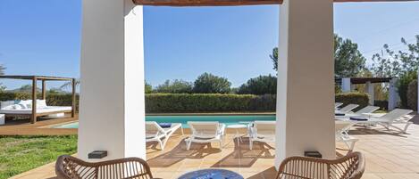Un espace extérieur abrité avec deux fauteuils en osier et une table, une piscine et des chaises longues en arrière-plan.