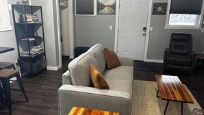 Ein Wohnzimmer mit grauem Sofa, zwei Holztischen, einem schwarzen Ledersessel, einem Bücherregal und einer Tür mit einem Schild.