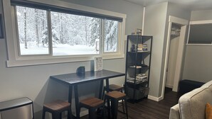 Ein gemütliches Zimmer mit Esstisch und Stühlen, einem Bücherregal und Blick auf einen verschneiten Wald durch ein großes Fenster.
