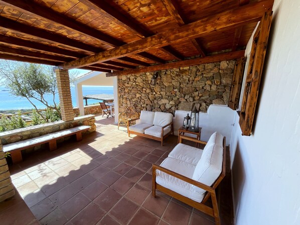 A covered patio with stone walls, wooden furniture, and a view of the sea. - DOMUS SUN: Sea and Sardinian Relaxation! (VILLASIMIUS CAMPULONGU)