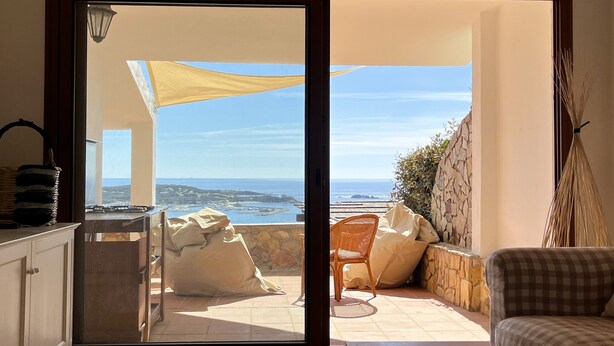 A room with a sliding glass door leading to a patio with a view of the sea. There is a wicker basket, a chair, and a checkered sofa inside the room.