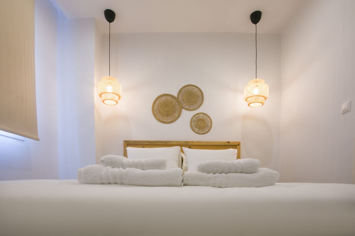 Un lit bien fait, avec du linge de lit et des serviettes blanches, deux lampes suspendues et trois éléments de décoration murale en fibres tressées.