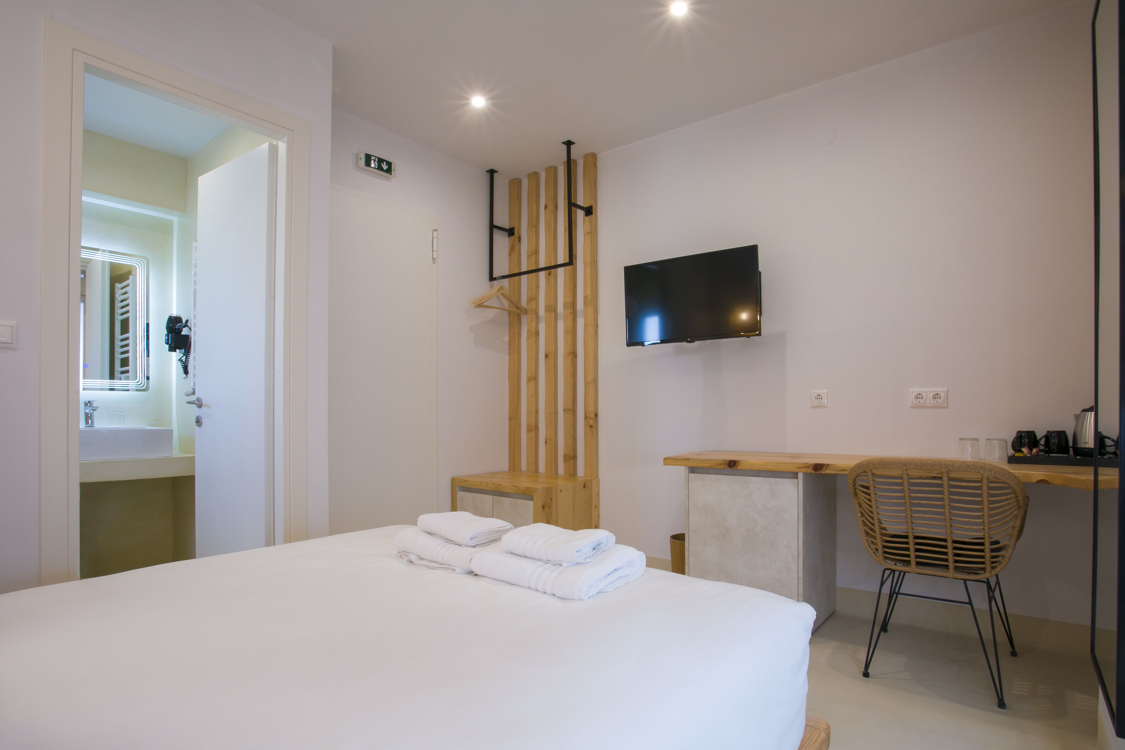 A hotel room with a bed, a TV mounted on the wall, a wooden chair, and a desk with a coffee maker.