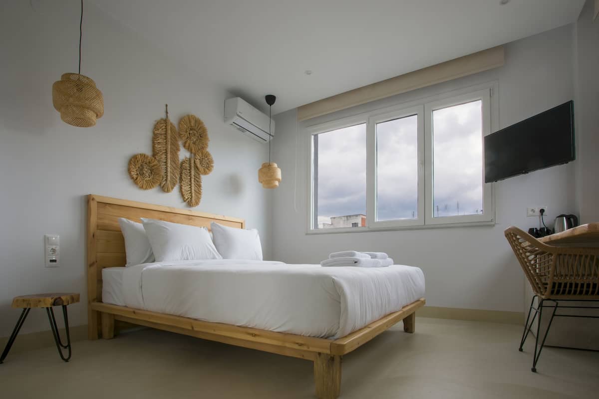Une chambre avec un lit en bois, une grande fenêtre, un téléviseur à écran plat et un fauteuil en osier.