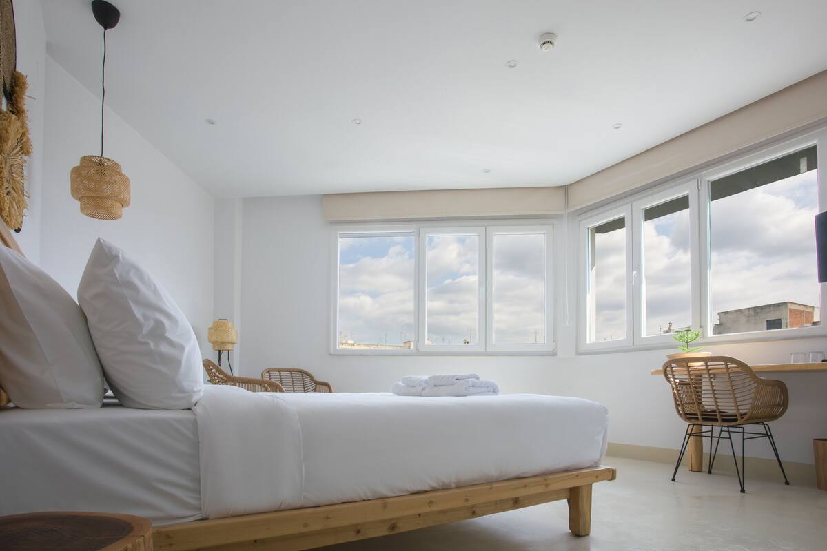 Une chambre à coucher moderne avec un lit, un banc en bois, une chaise en osier et un bureau avec une lampe.