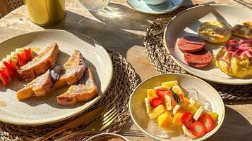 Un desayuno con tostadas, fruta, bollería, una taza de café y un florero con planta.