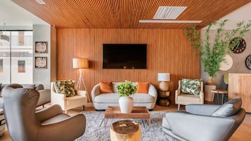 Uma sala de estar moderna com teto de ripas de madeira, uma TV de tela plana na parede e diversos assentos, incluindo um sofá cinza e poltronas.