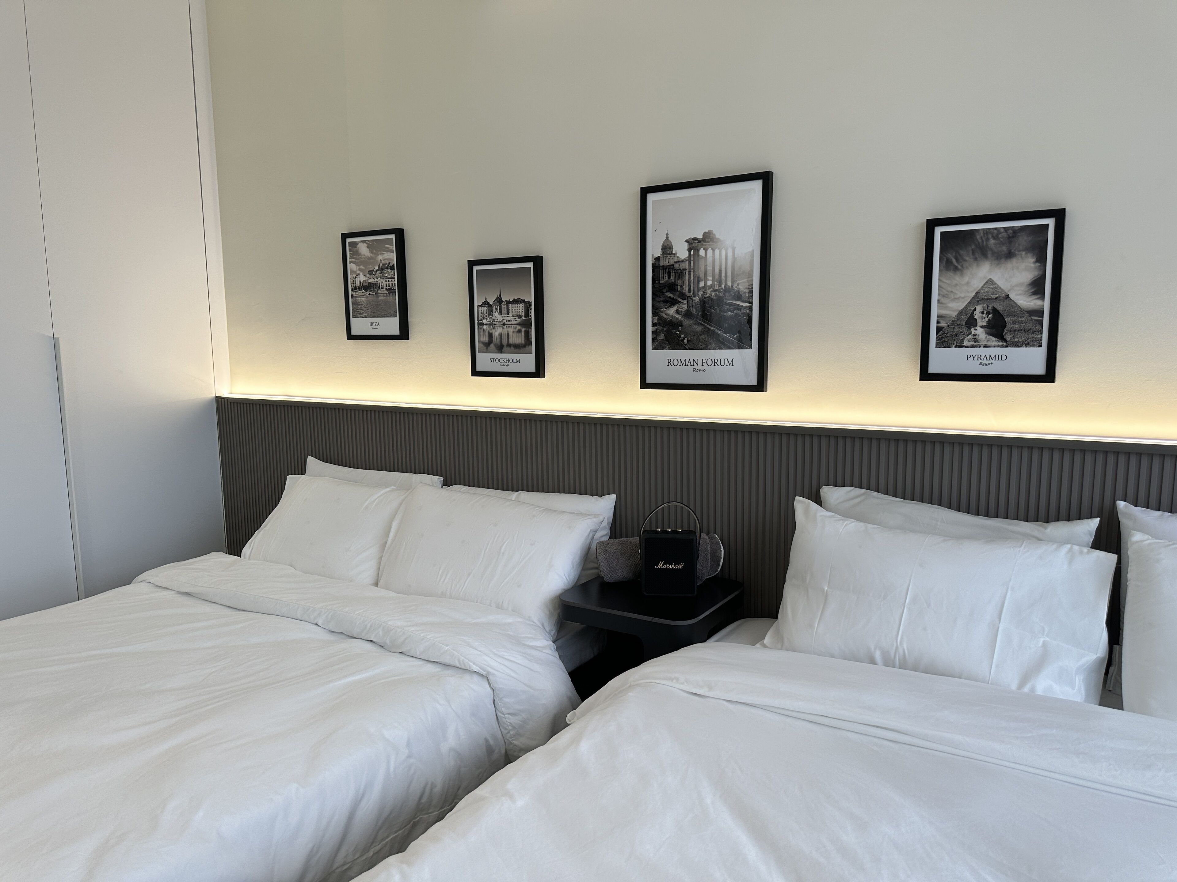 Ein Hotelzimmer mit zwei Betten, einem Nachttisch und gerahmten Bildern bekannter Sehenswürdigkeiten.