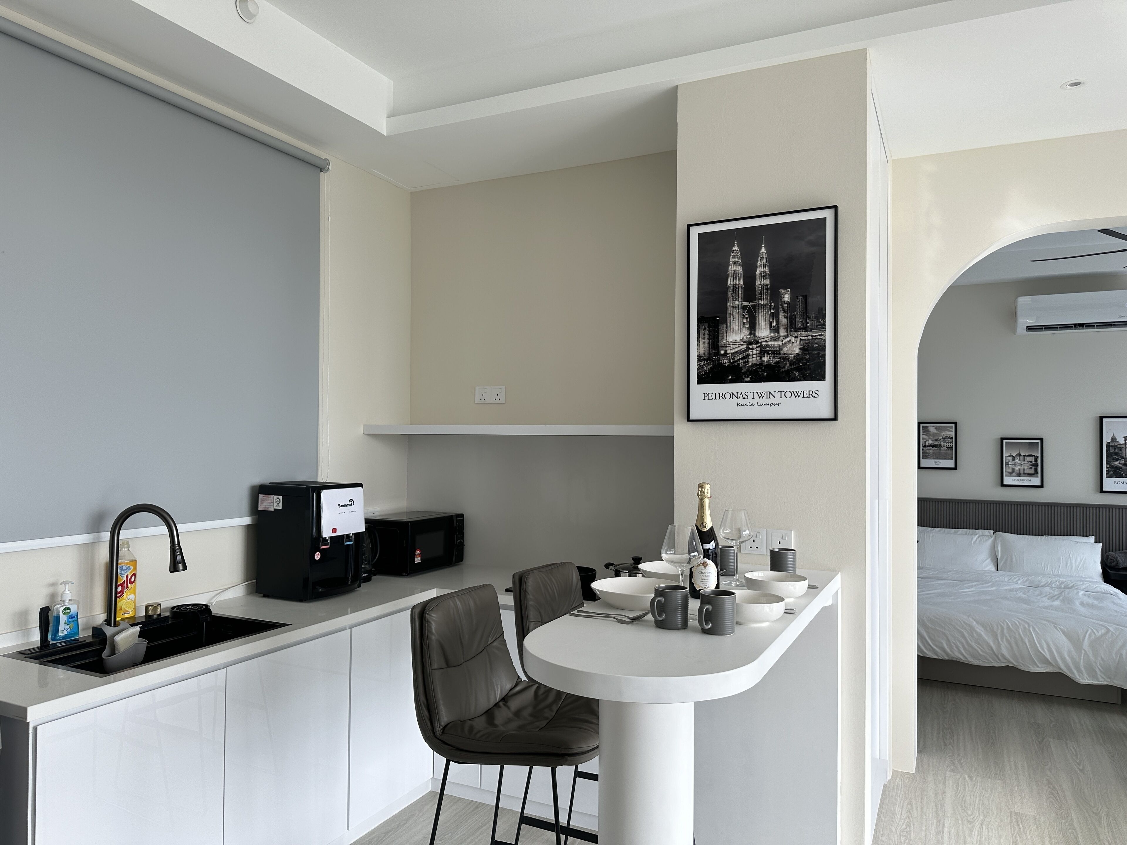Una cocina moderna con encimera blanca, microondas integrado y un lavamanos. Hay una zona de bar con una silla y una botella de champán. En la pared, se observa un cuadro con las Torres Petronas.