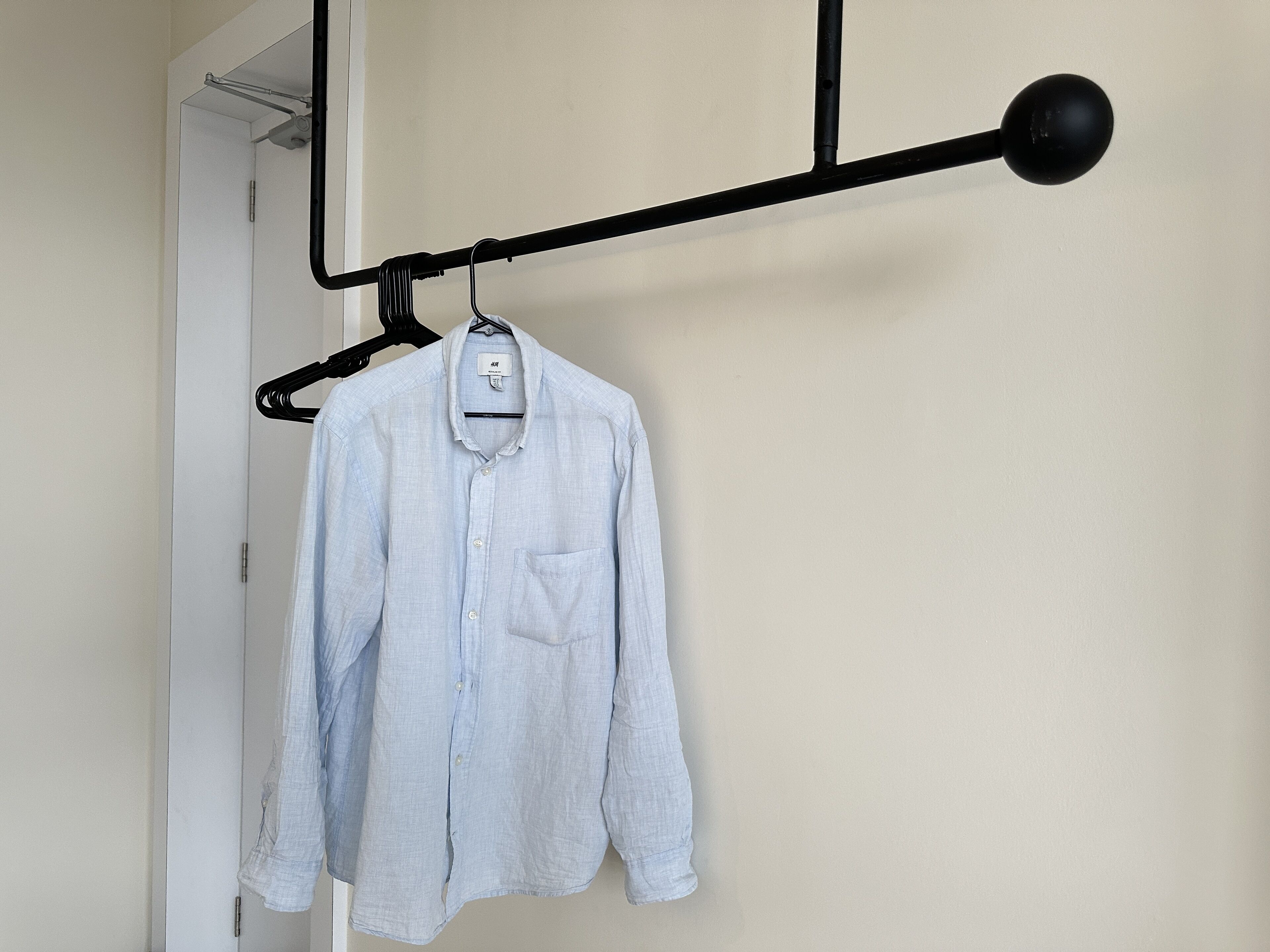A light blue button-up shirt hanging on a black hanger against a plain wall.