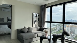 A modern living room with a grey sofa, a glass coffee table, and a large window offering a city view.