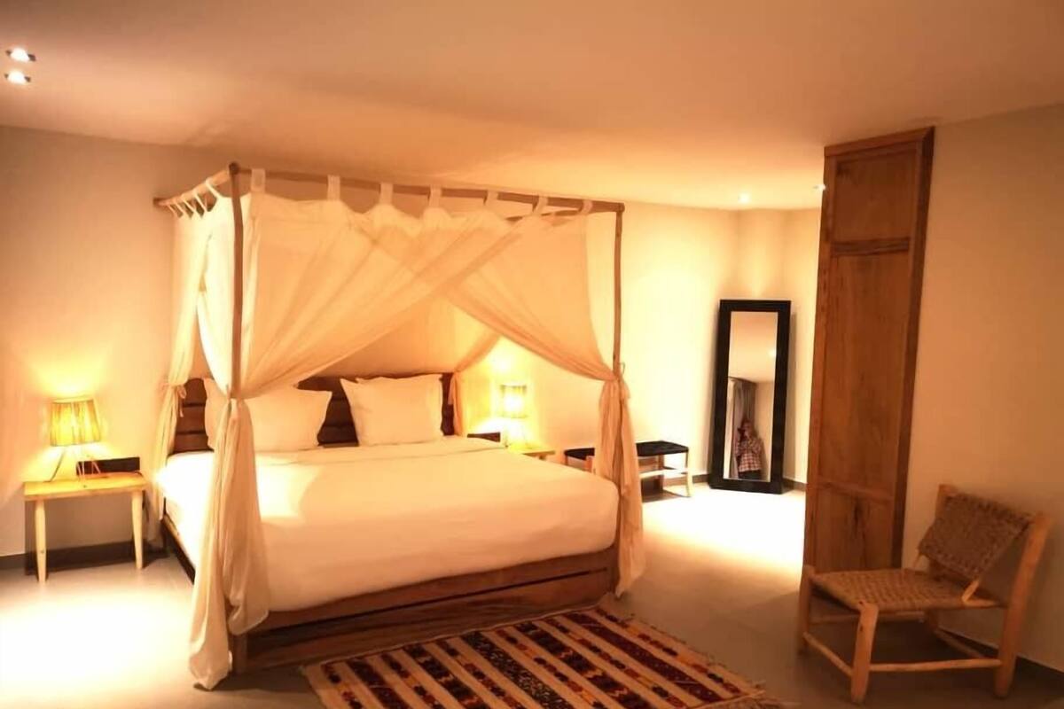 Ein Bett mit Baldachin und hölzernem Gestell, ausgestattet mit weißer Bettwäsche, ein Nachttisch aus Holz mit einer Lampe, ein gewebter Teppich und ein Holzstuhl.