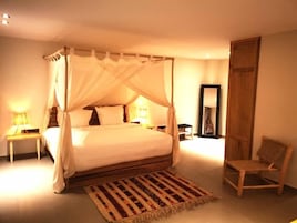 A four-poster canopy bed with a wooden frame and white bedding, a wooden nightstand with a lamp, a woven rug, and a wooden chair.