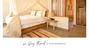 A bedroom with a four-poster bed, a bedside table with a lamp, and a patterned rug. - Les Goréennes Suites (Mbour)