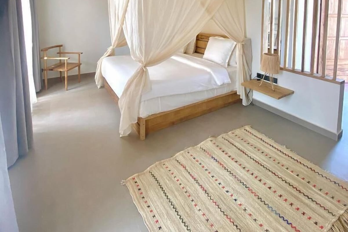 A bedroom with a canopy bed, a small wooden chair, a bedside table with a lamp, and a patterned rug.