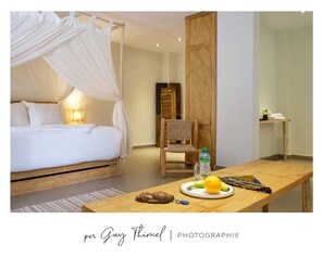 A bedroom with a canopy bed, a wooden chair, a table with a plate of fruit and a bottle, and a nightstand with a lamp. - Les Goréennes Suites (Mbour)