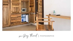 A wooden cabinet with open doors, a microwave, and a small table with a bottle and remote control. - Les Goréennes Suites (Mbour)