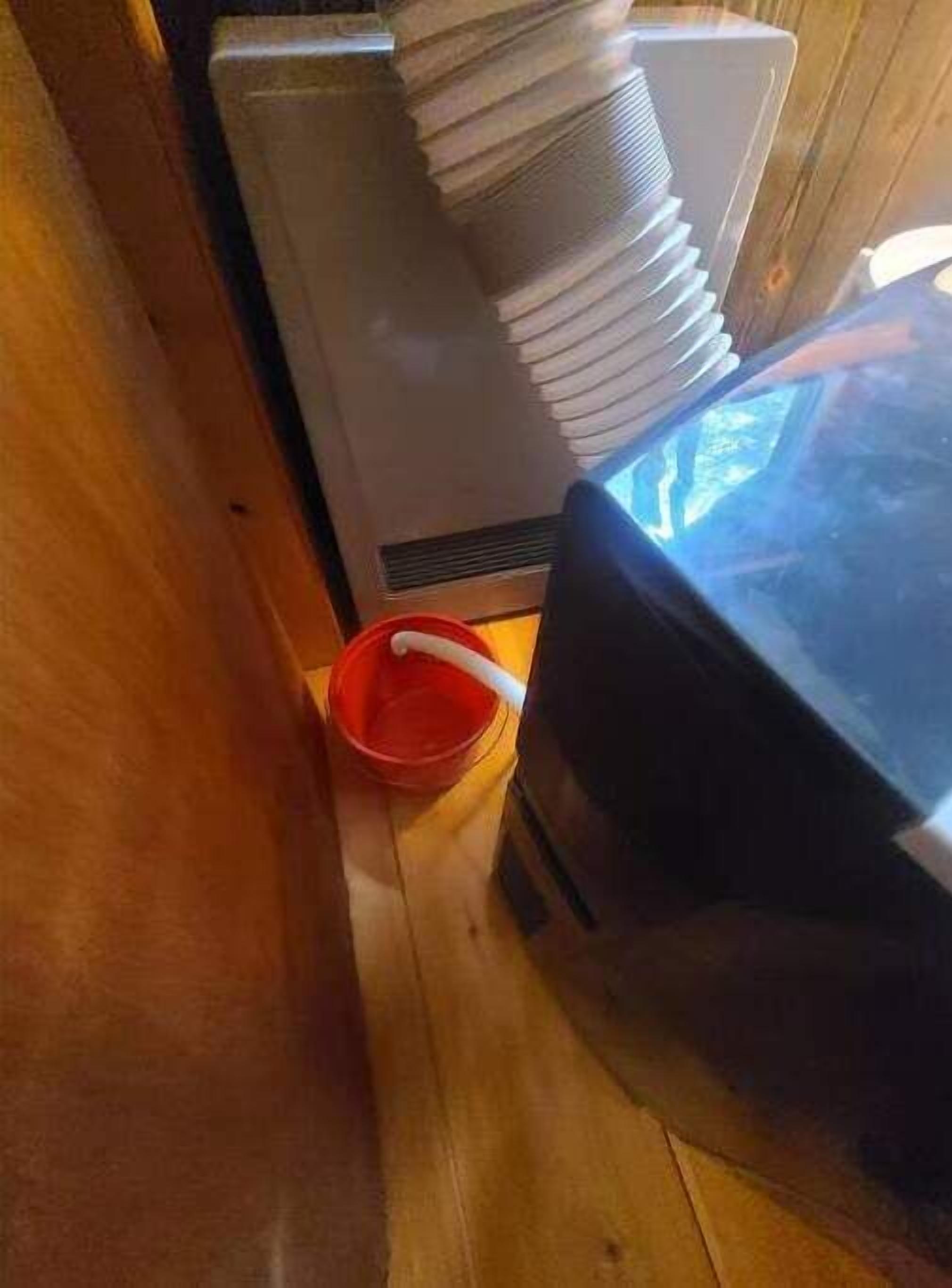 A compact kitchen with a microwave, a red bucket, and a stack of plates on a wooden cabinet.