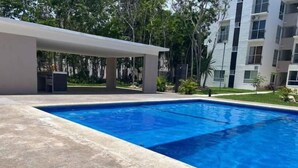 Una piscina rectangular con una solana empavedada, rodeada de vegetación y un edificio moderno al fondo.