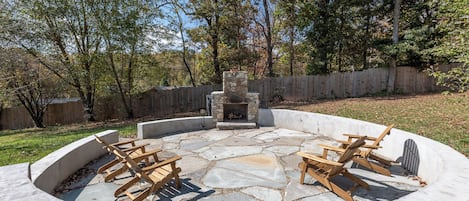 A circular stone fire pit with wooden chairs and a stone fireplace in the center, surrounded by a wooden fence and trees.