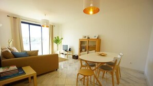 A modern living room with a sofa, a dining table with chairs, a bookshelf, and a TV. - Alojamento Santa Maria B4 1E (Tavira)