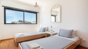 A modern bedroom with two single beds, a large window, a bedside table with a lamp, and a mirror on the wall.