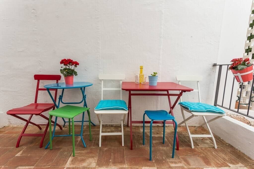 A small outdoor seating area with a red table and chairs, a blue table with a potted plant, and a white table with a yellow bottle.