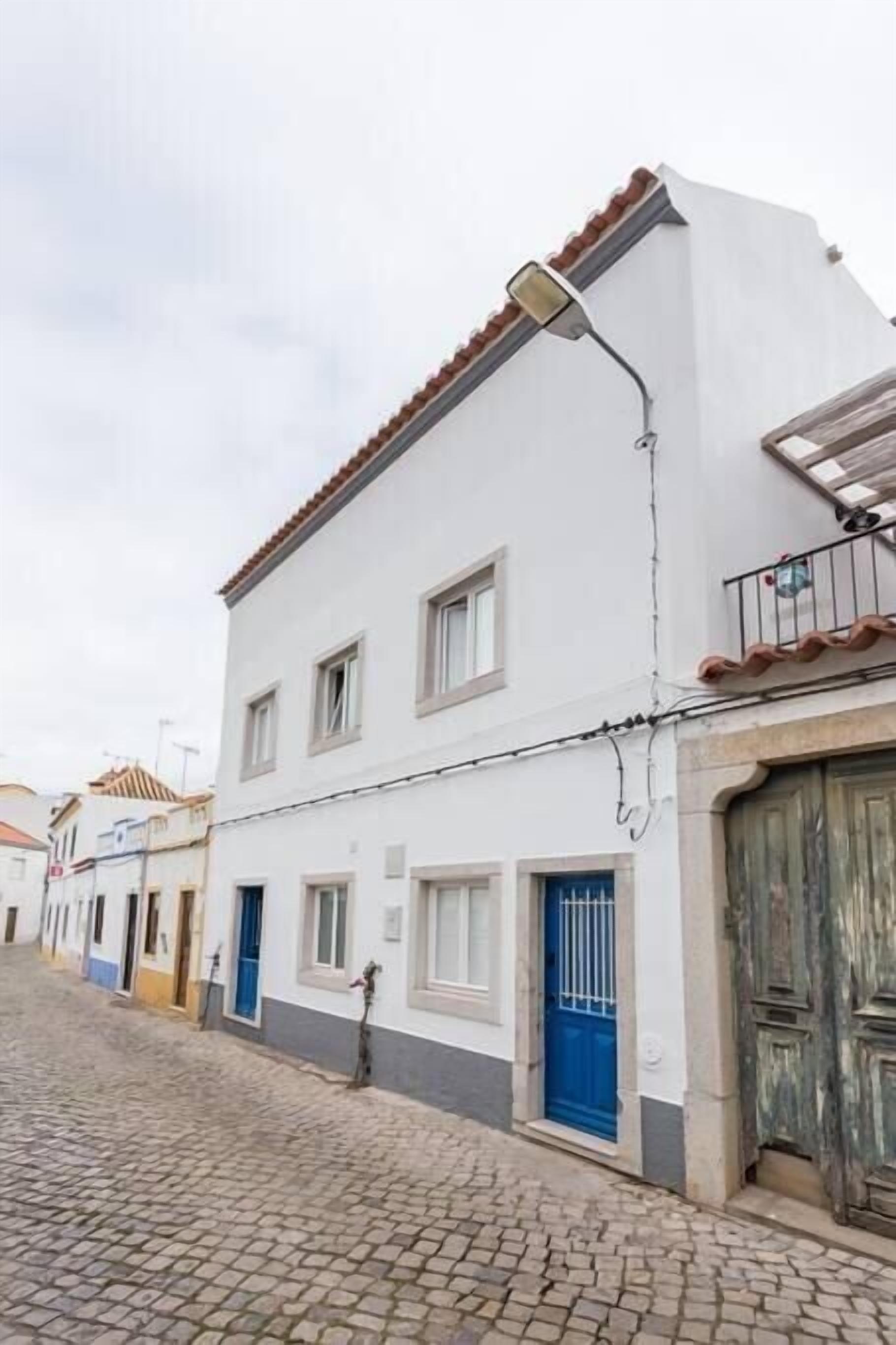 Une ruelle étroite pavée, bordée d’un bâtiment blanc avec des portes bleues et un toit recouvert de tuiles.