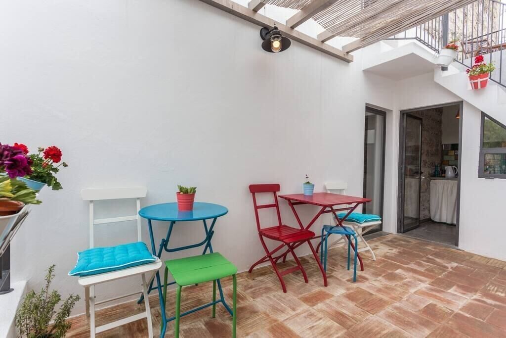 Un patio con pared blanca, techo de madera y suelo de baldosas. Hay dos mesas y cuatro sillas, una azul y una roja.