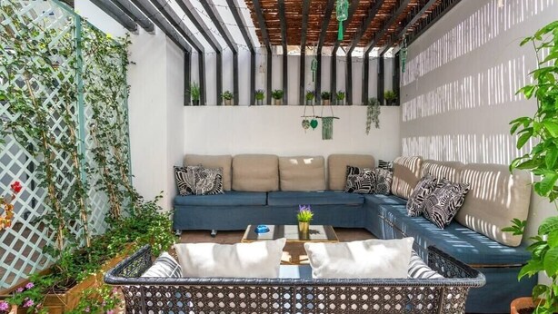 Un rincón exterior acogedor con un sofá azul, cojines estampados y una mesa de mimbre.