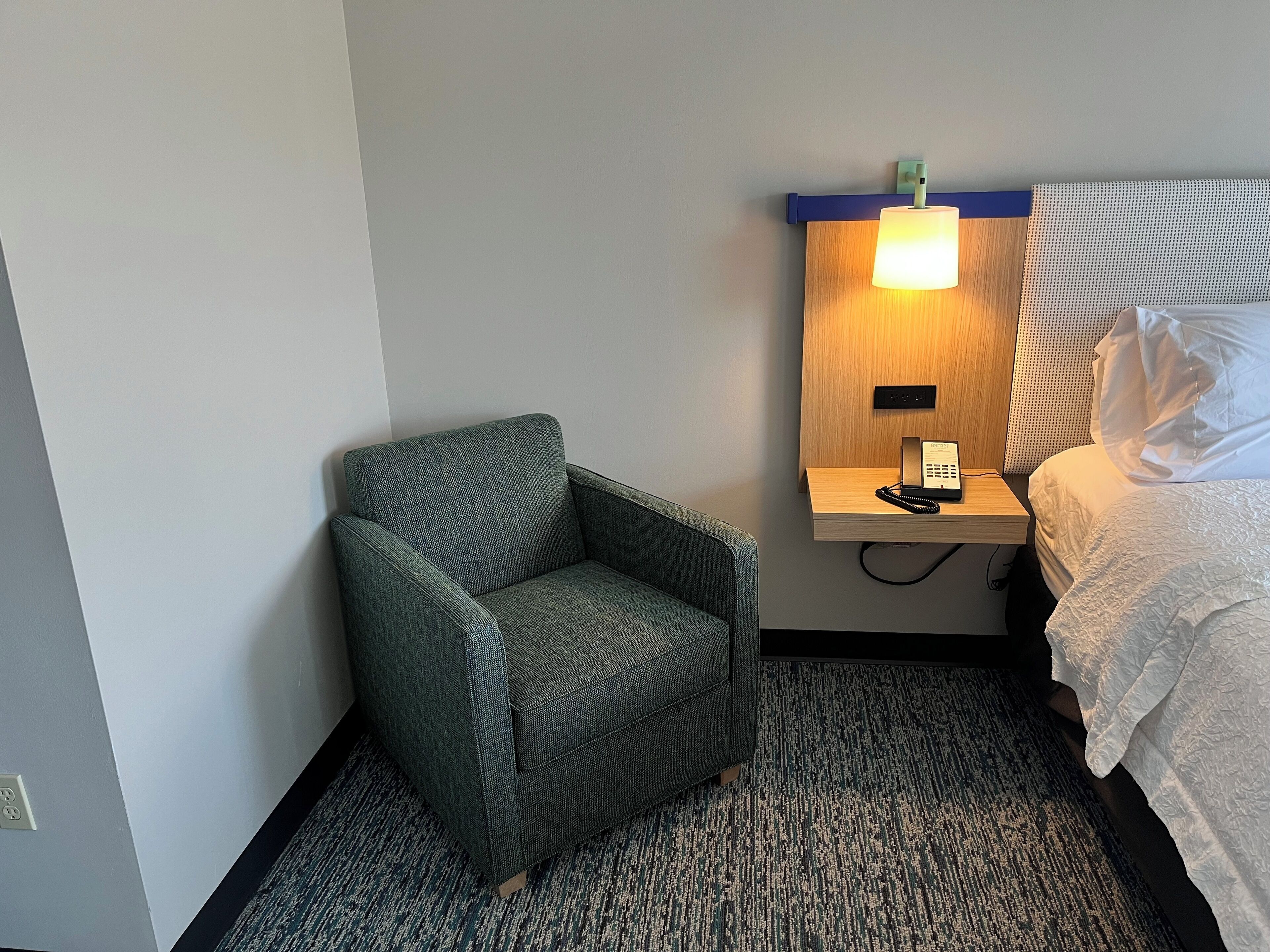 A hotel room with a grey armchair, a bedside table with a lamp, and a bed with white bedding.