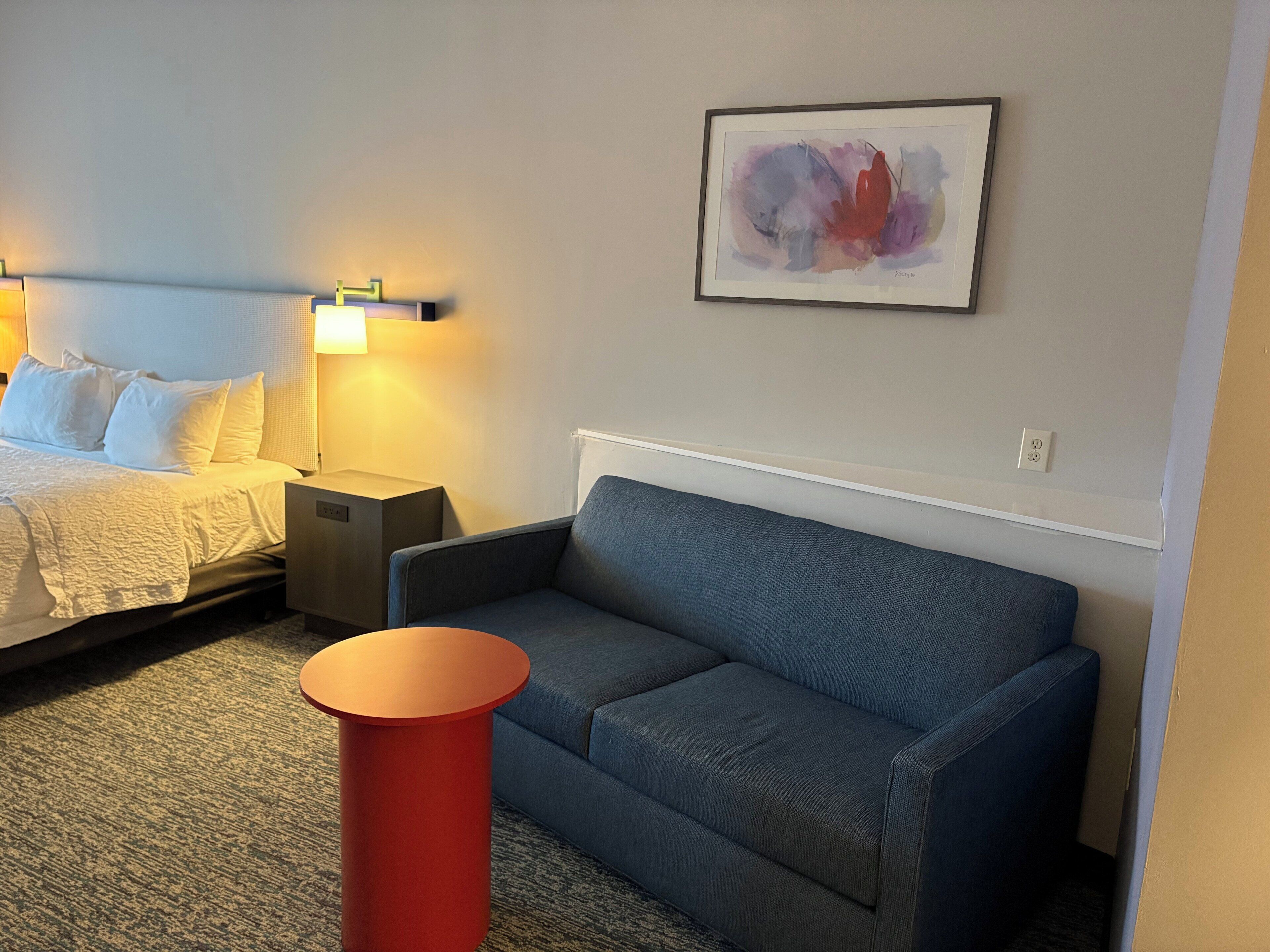 A hotel room with a bed, a blue sofa, a red side table, and a framed abstract painting on the wall.