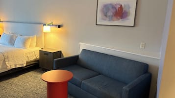 A hotel room with a bed, a blue sofa, a red side table, and a framed abstract painting on the wall.