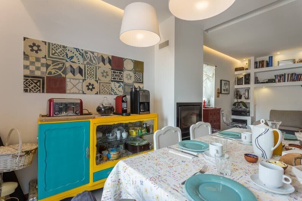 Un comedor con una mesa preparada para cuatro personas, un armario amarillo y azul con utensilios de cocina, y una pared con un colorido diseño de azulejos.