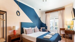 A hotel room with a bed, bedside table, lamp, chair, and a view of the city through the window.
