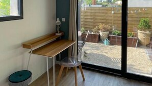 Una habitación con un escritorio de madera, una silla, un taburete pequeño y un amplio ventanal corredera que da a un jardín con plantas en macetas.