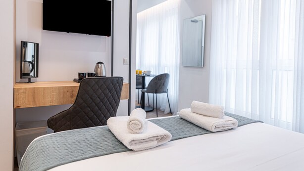 A hotel room with a bed, a desk with a computer, a chair, and a TV mounted on the wall.