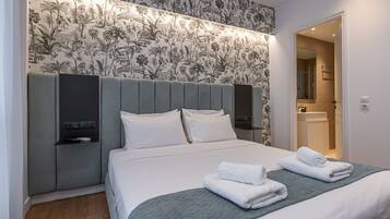 Una cama bien hecha con sábanas blancas y una manta gris, un cabecero con diseño de papel tapiz floral, y un baño visible a través de una puerta abierta.