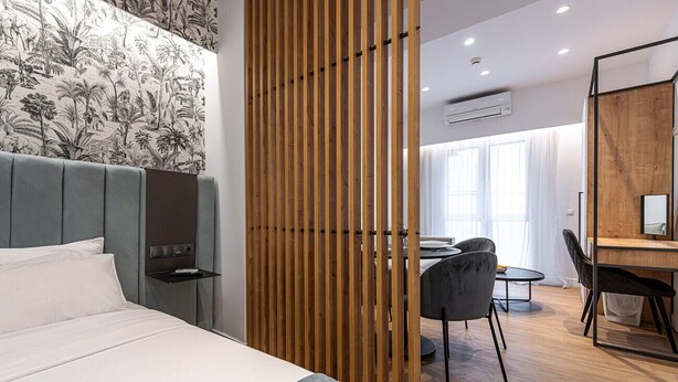 Una habitación de hotel moderna con una cama grande, un separador de madera, un escritorio con silla y un ventanal amplio.