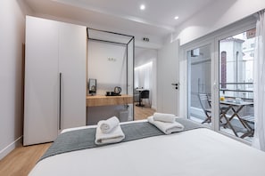 A modern hotel room with a large bed, a wooden side table, a mirror, and a balcony with a table and chairs. - Kriel Suites by LIV Homes 101 (Athens)
