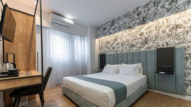 A hotel room with a bed, a desk with a chair, a television, and a wall with a floral pattern. - Kriel Suites by LIV Homes 203 (Athens)