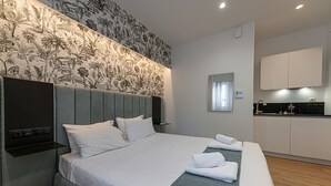 A modern hotel room with a large bed, a built-in headboard, a small kitchenette, and a decorative wall with a floral pattern. - Kriel Suites by LIV Homes 203 (Athens)