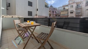 A balcony with a table and chairs, a glass of orange juice, and a view of surrounding buildings. - Kriel Suites by LIV Homes 203 (Athens)