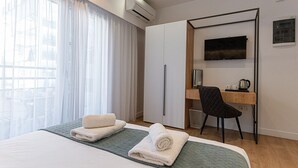 A hotel room with a bed, a desk, a chair, a TV, and an air conditioning unit. - Kriel Suites by LIV Homes 202 (Athens)